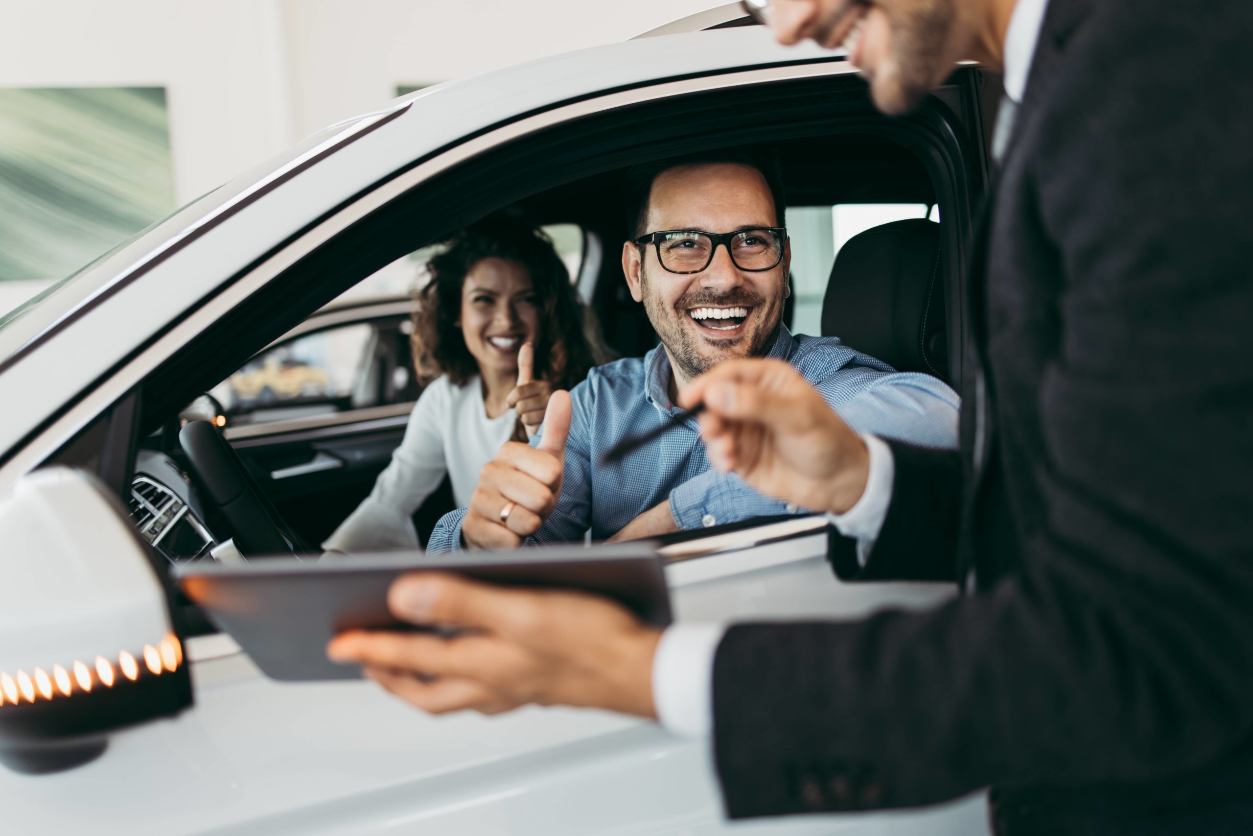 Middle aged couple sitting in their new car giving the car salesman a thumbs up.