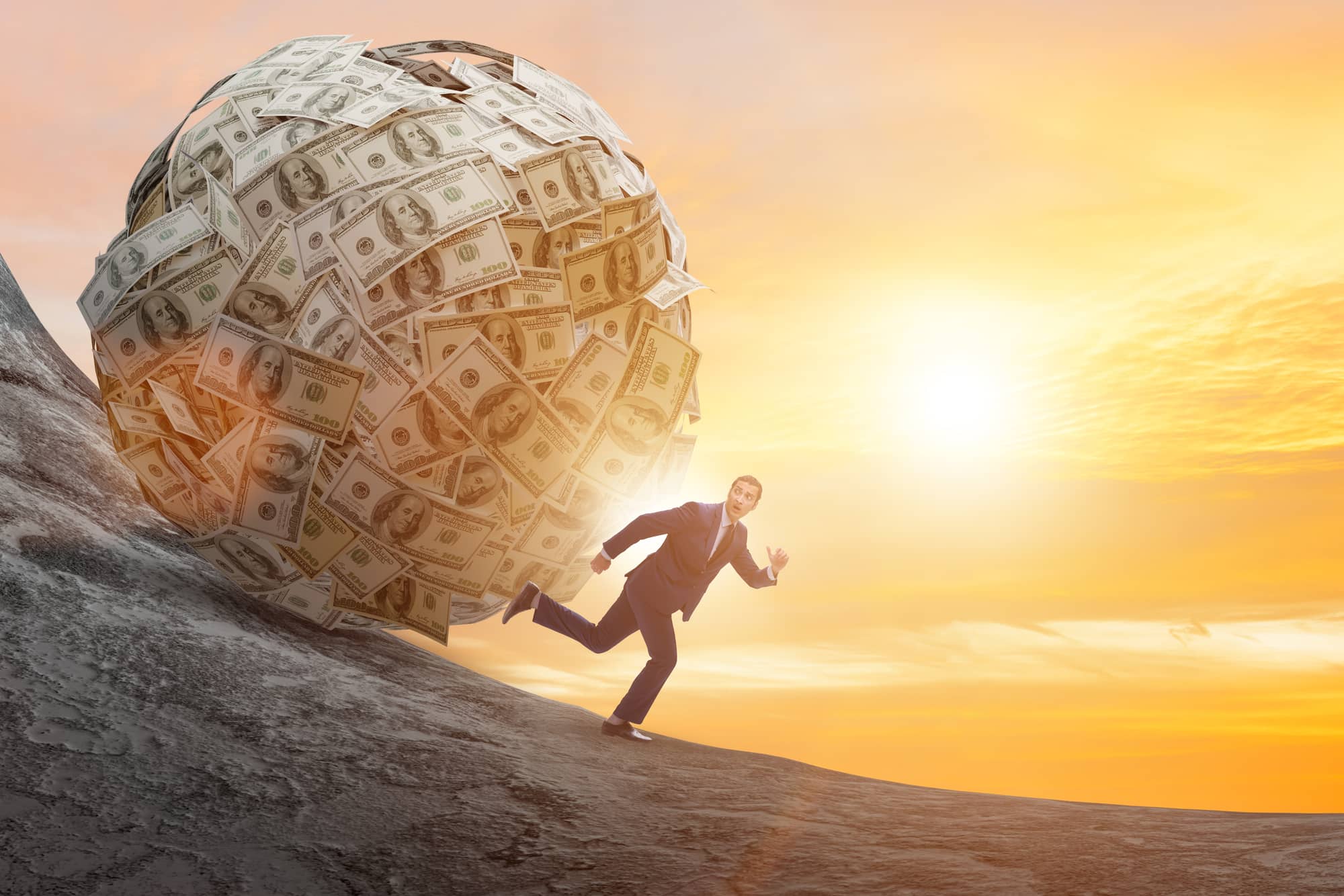 Man running away from a large ball of money rolling downhill.